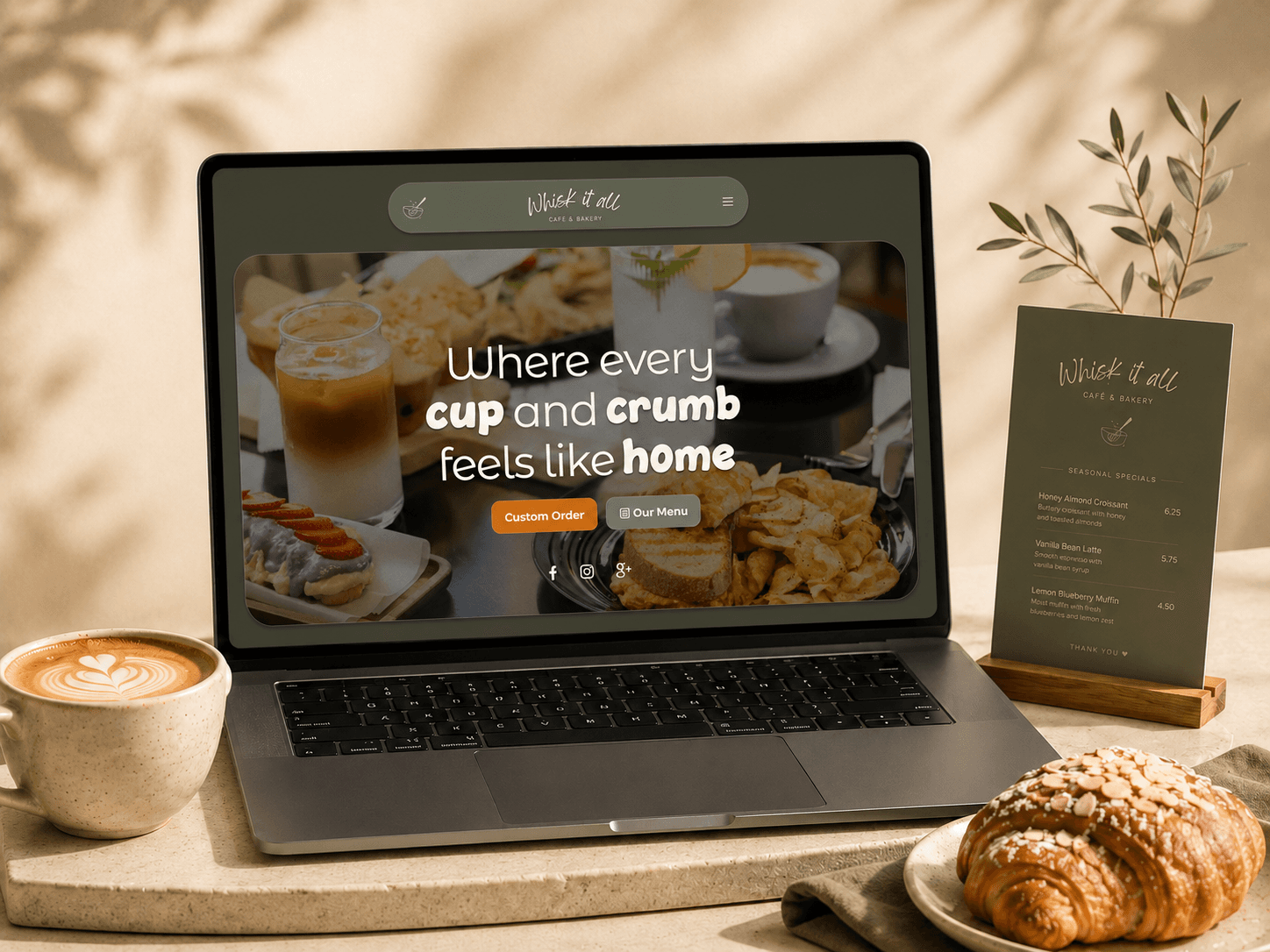 The Whisk It All homepage open on a laptop at a café table, beside a latte, a croissant, and a printed specials card.
